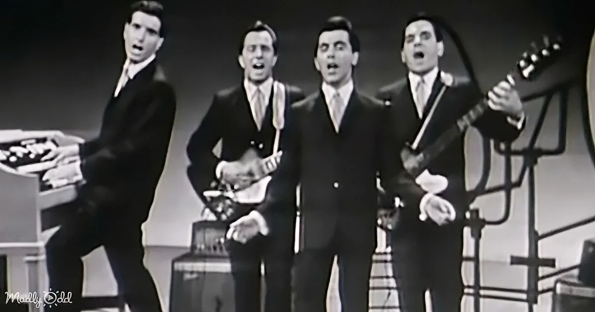 Four men in suits perform on stage, one playing guitar, others singing, with a vintage aesthetic.