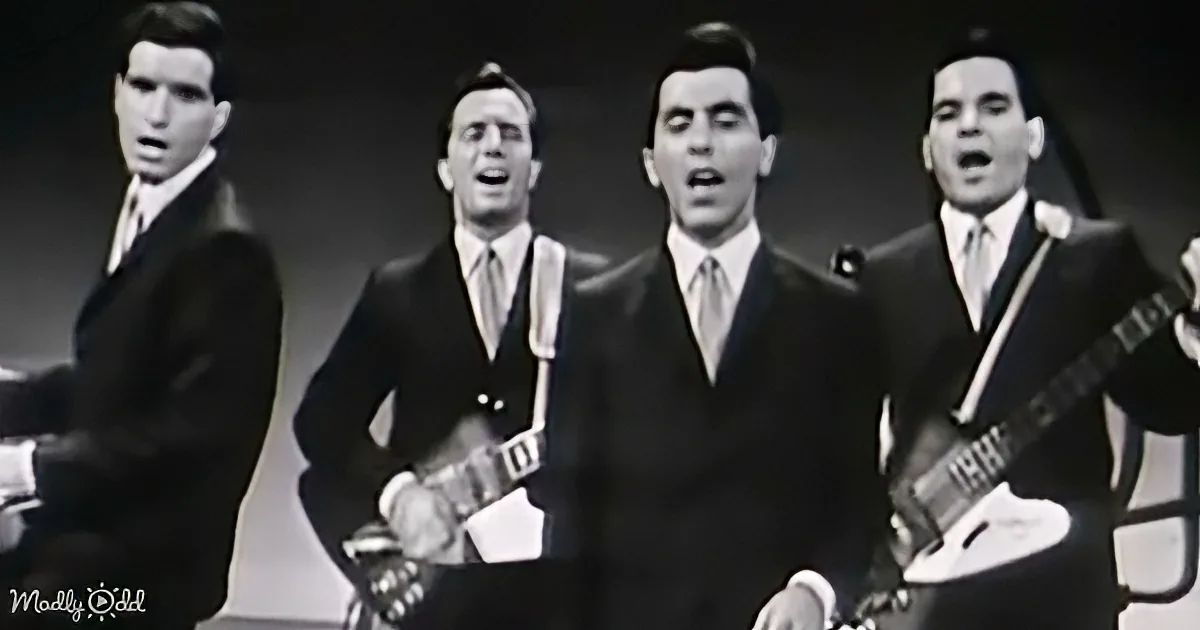 Four men in suits play guitars and sing in a vintage black-and-white setting.