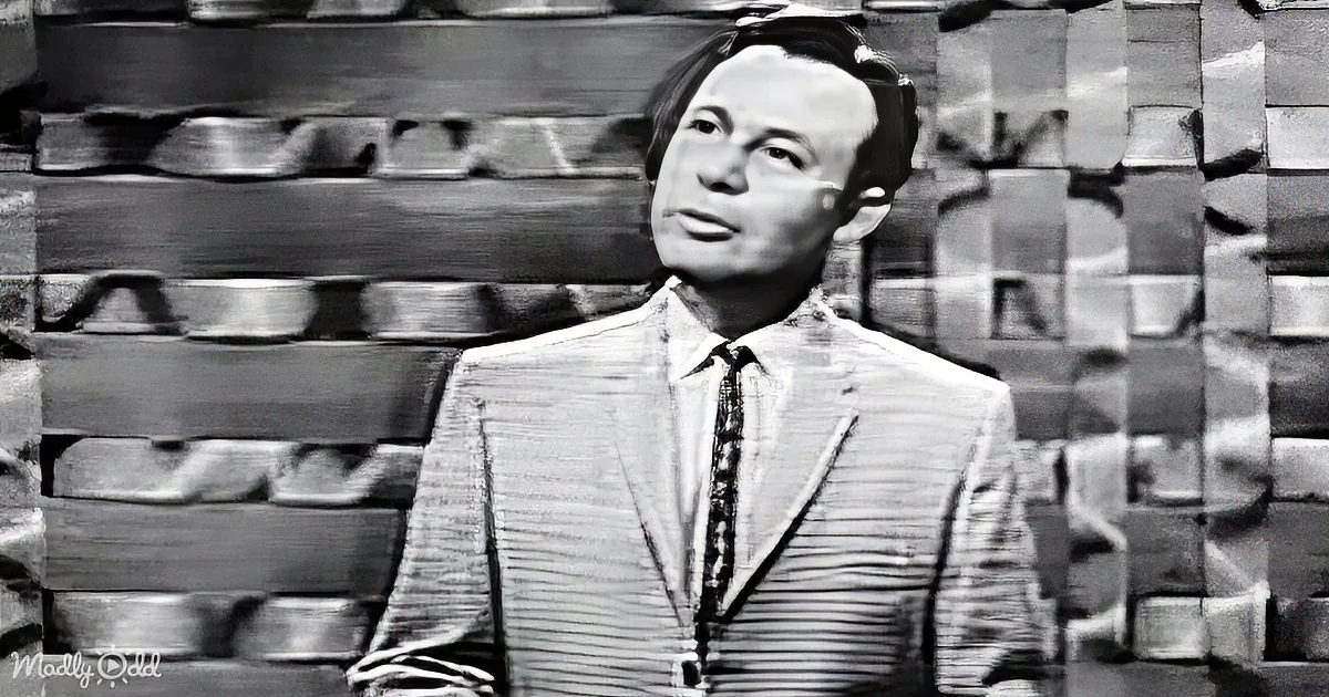 A man in a suit and tie stands against a blurred brick wall backdrop.