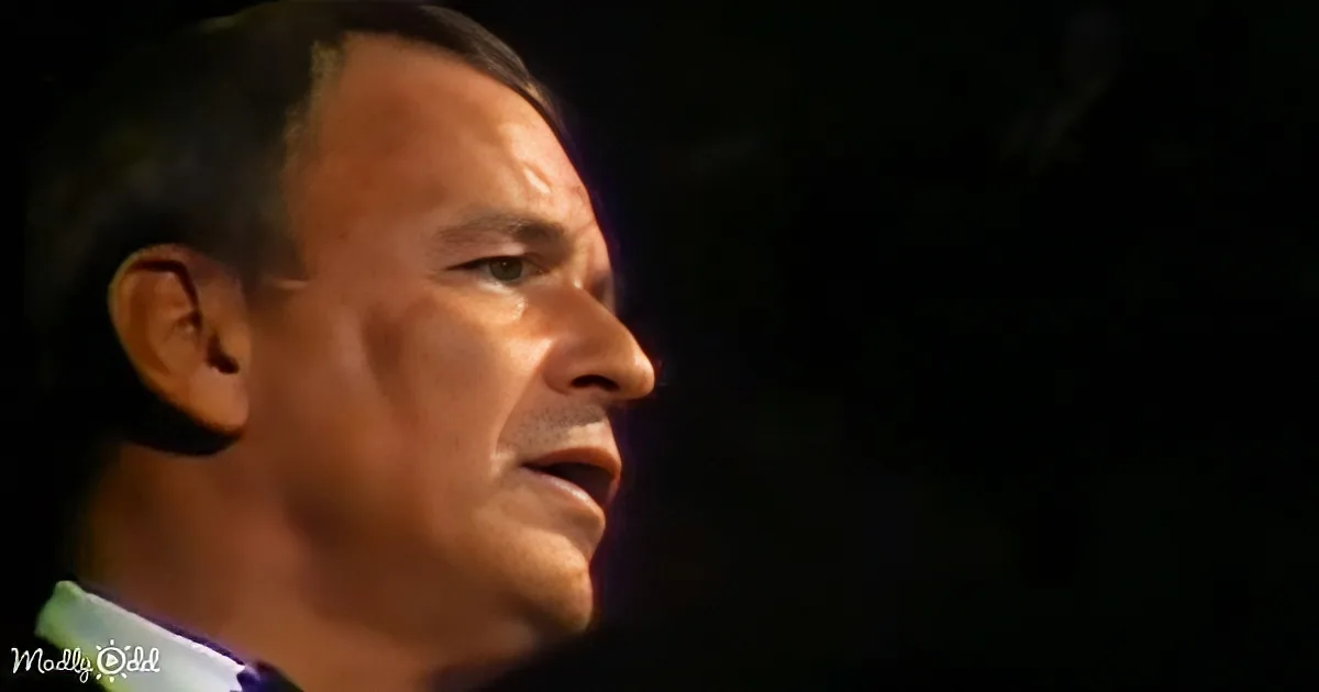A man in a suit and tie is speaking, his face partially illuminated against a dark background.