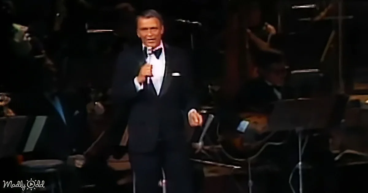 A man in a tuxedo sings into a microphone on stage with an orchestra in the background.