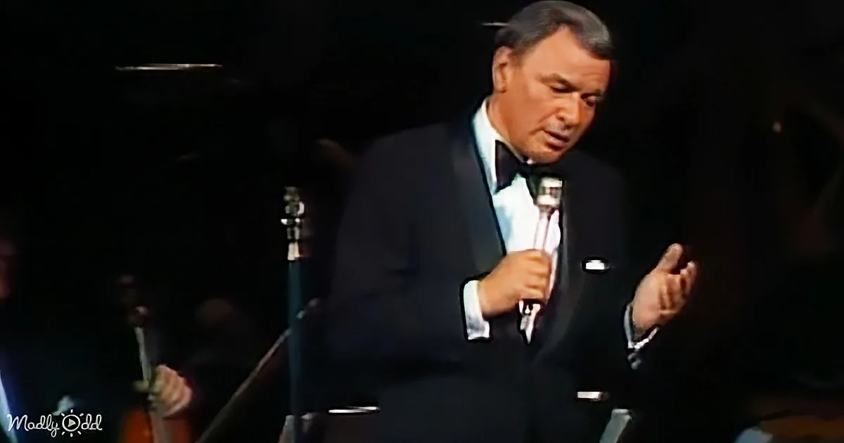 A man in a tuxedo sings into a microphone, gesturing with his hands.