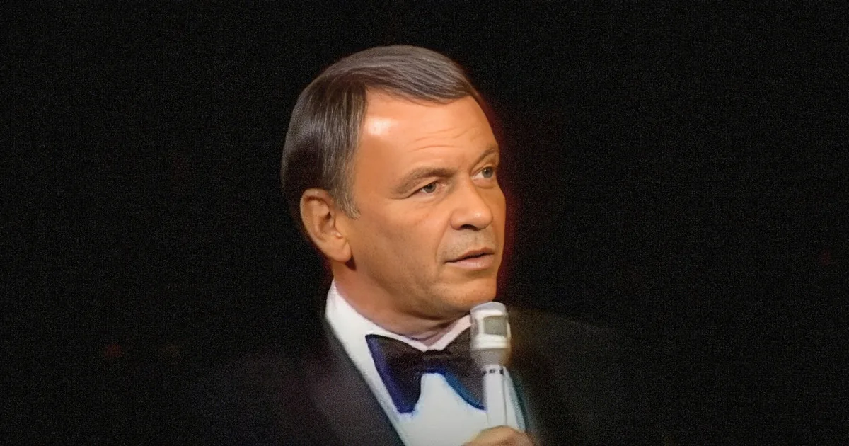 A man in a suit and bow tie holds a microphone, looking upwards with a focused expression.