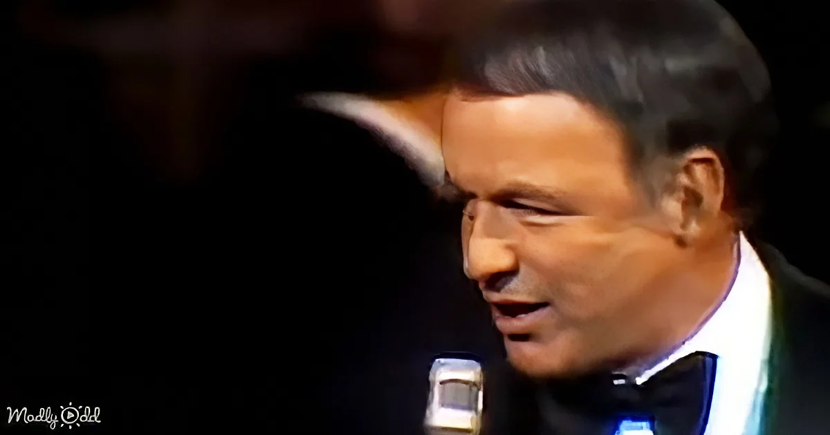A man in a suit and bow tie sings into a microphone, his face partially illuminated.