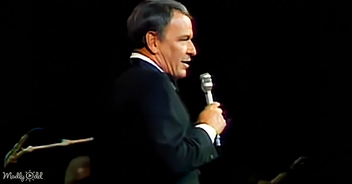A man in a suit holds a microphone, smiling, with a dark background.