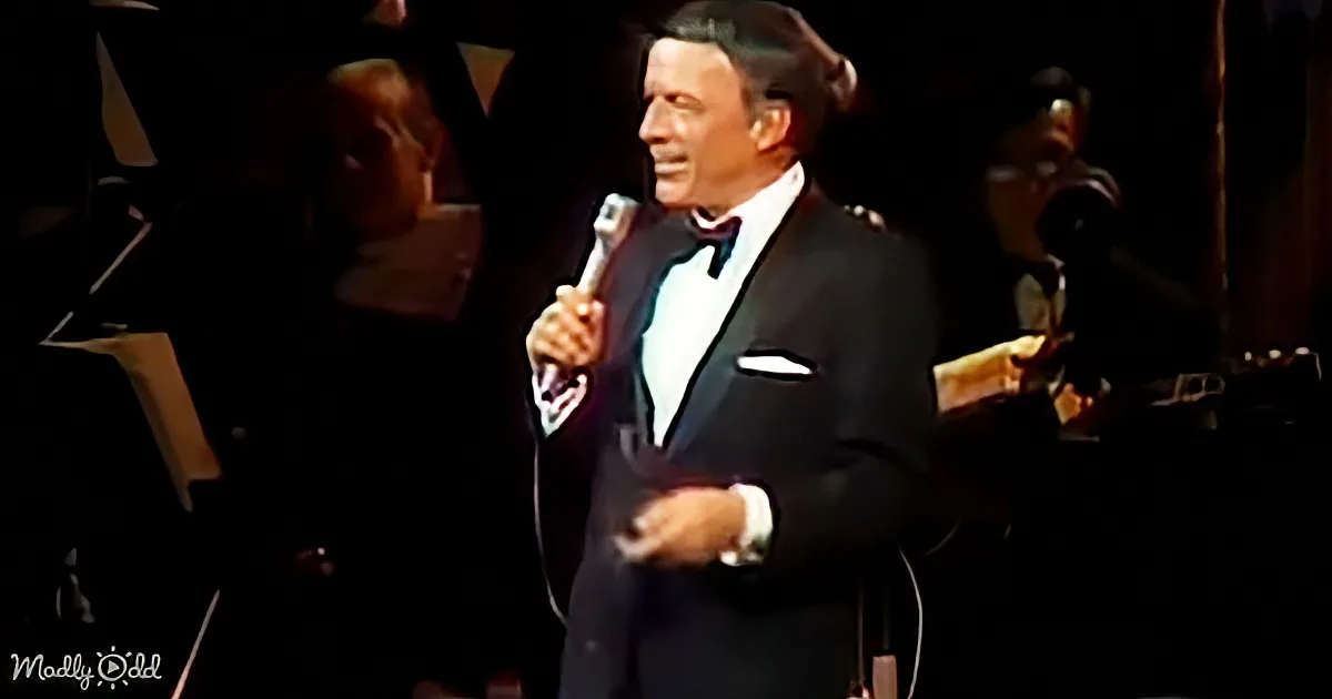 A man in a tuxedo sings into a microphone on stage with an orchestra in the background.