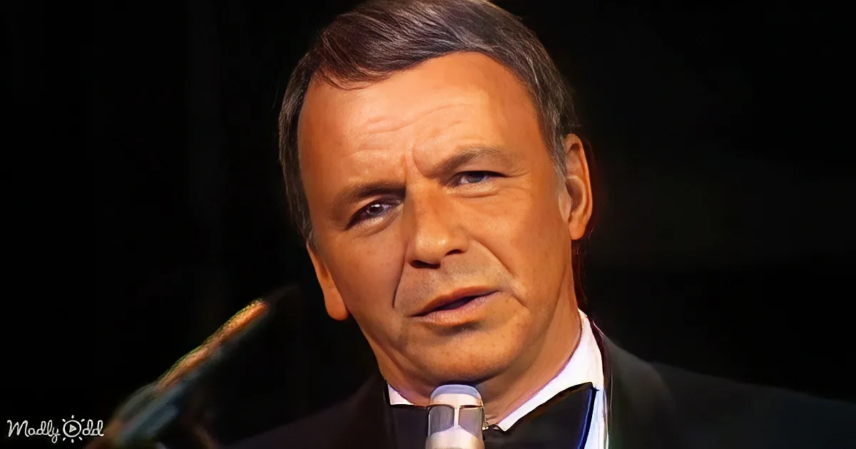A man in a suit and bow tie speaks into a microphone against a dark background.