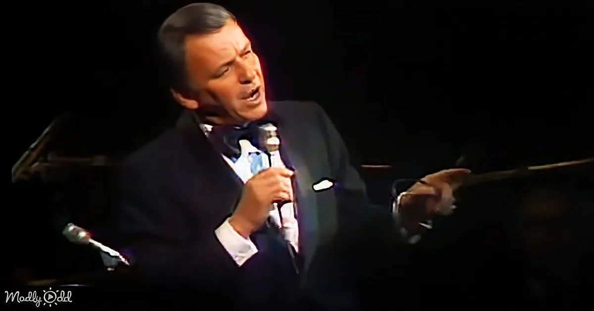 A man in a suit and bow tie sings into a microphone on stage, gesturing with his hand.