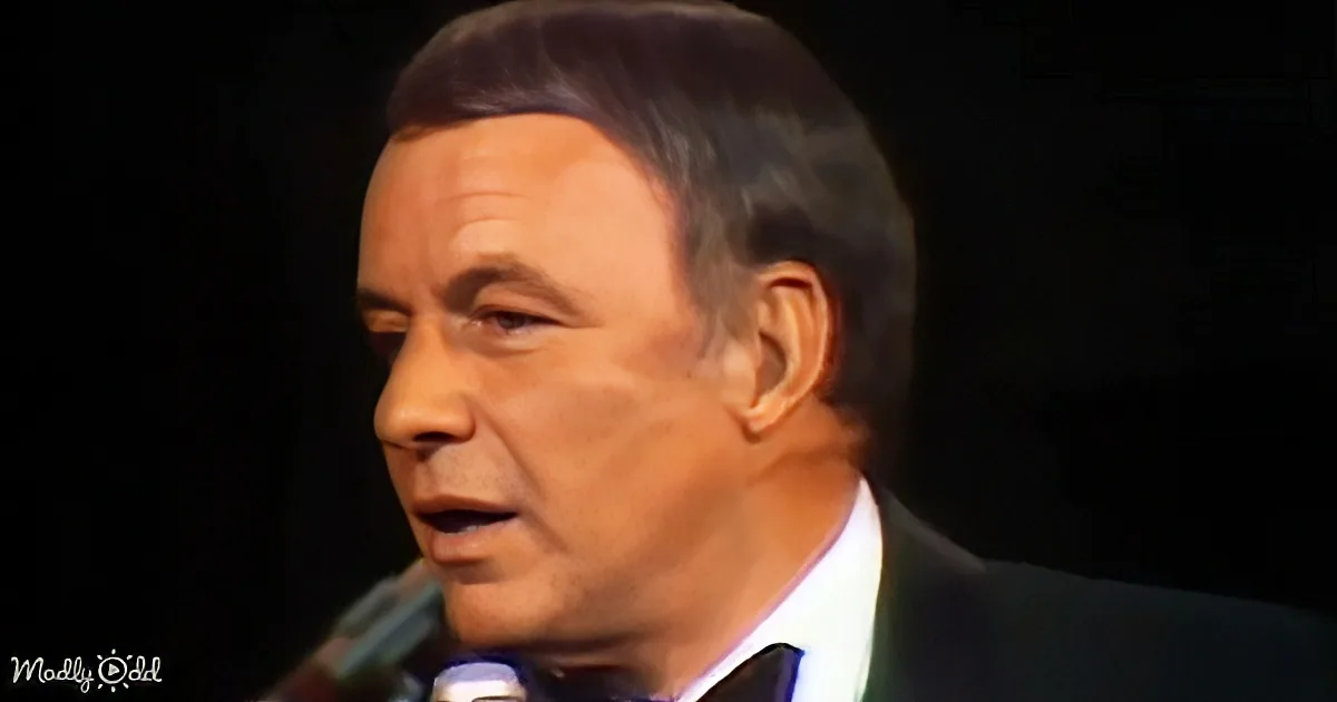 A man in a suit and tie is speaking into a microphone against a dark background.