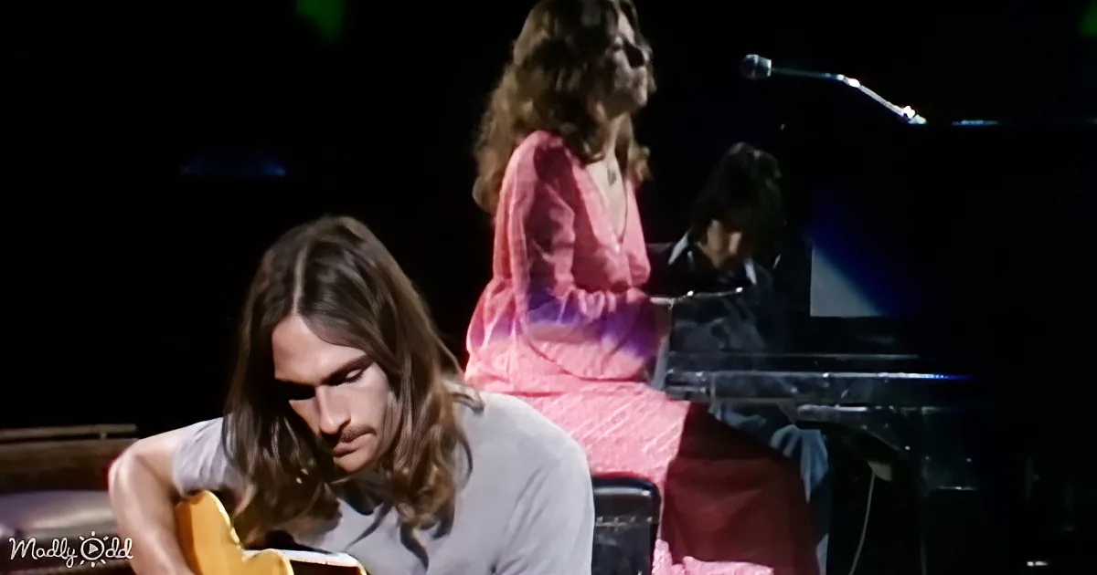 A man with long hair plays guitar while a woman in a pink dress sings and another person plays piano in the background.