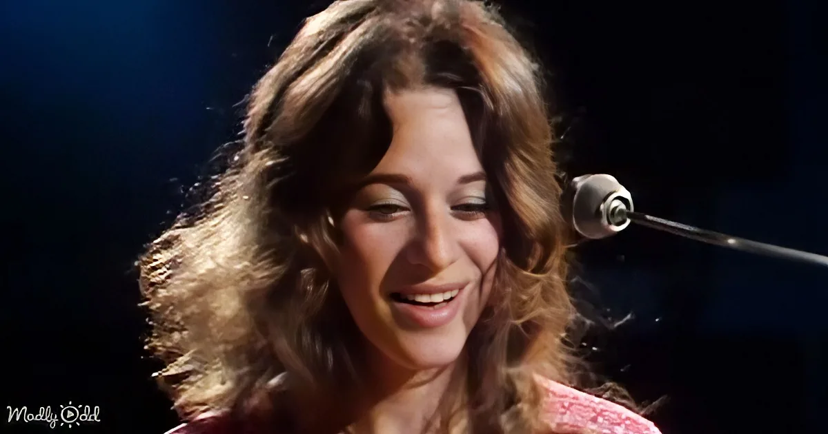 A woman with curly hair smiles near a microphone, wearing a pink top, with a dark background.