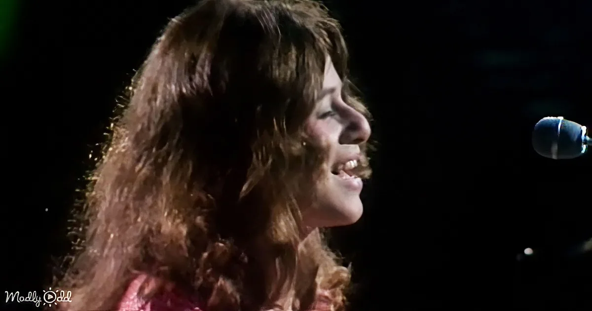A woman with long, curly brown hair smiles while singing into a microphone.