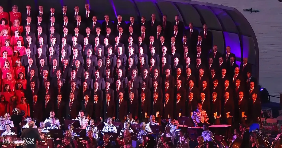 A large choir in formal attire stands in rows, accompanied by an orchestra on stage.