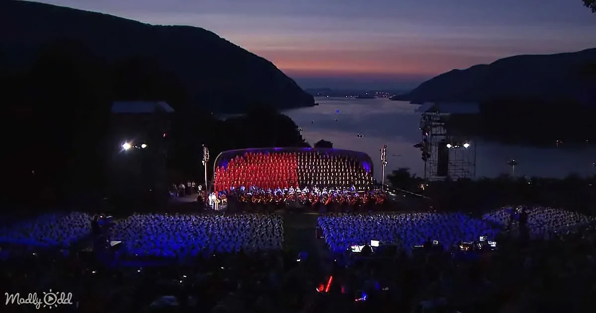 A large crowd gathers at a nighttime outdoor concert with a mountainous backdrop and a glowing stage.