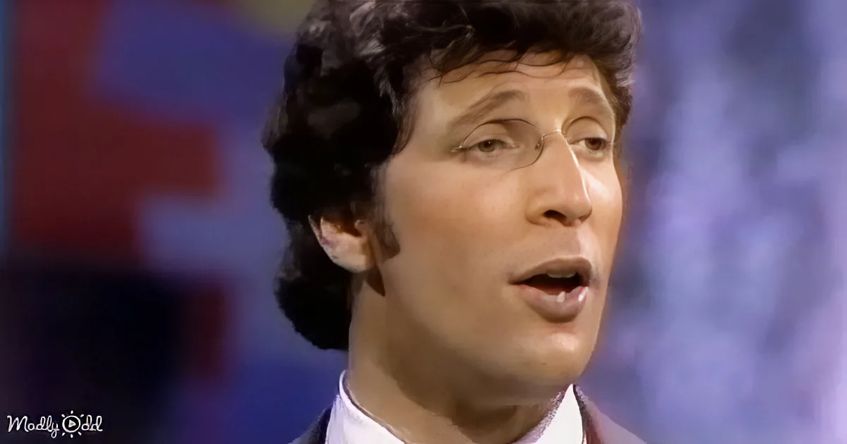 A man with curly hair, wearing a white shirt, appears to be speaking or singing.