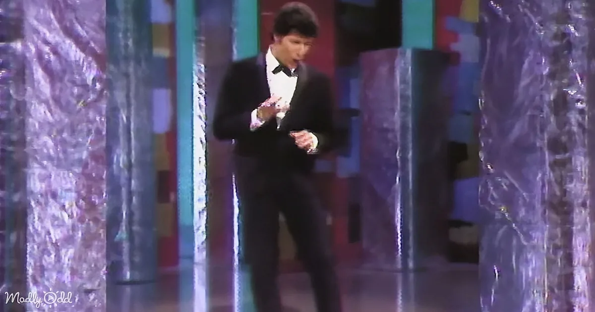 A man in a tuxedo stands on a stage with colorful, reflective columns behind him.