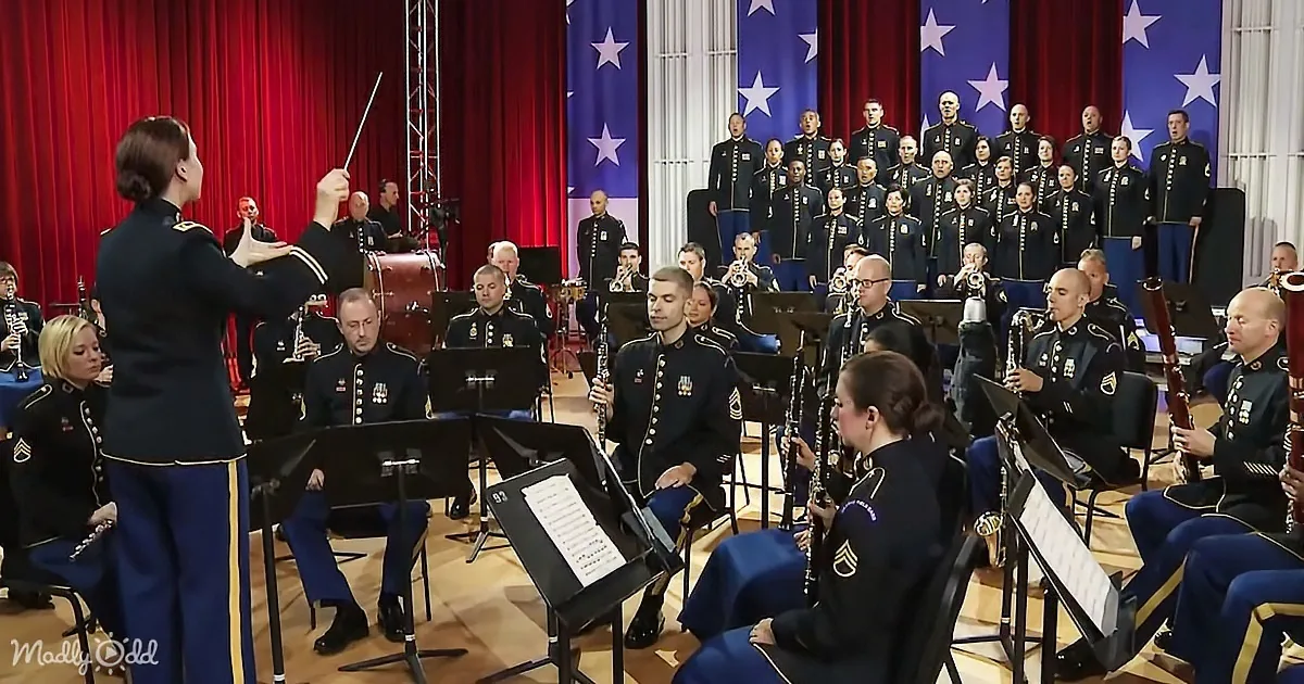 A conductor leads a military band and choir in uniform, with American flags in the background.