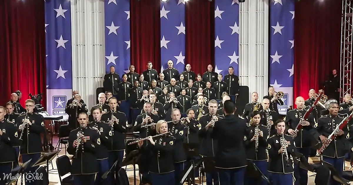A military band in uniform plays instruments on stage with American flags and red curtains in the background.