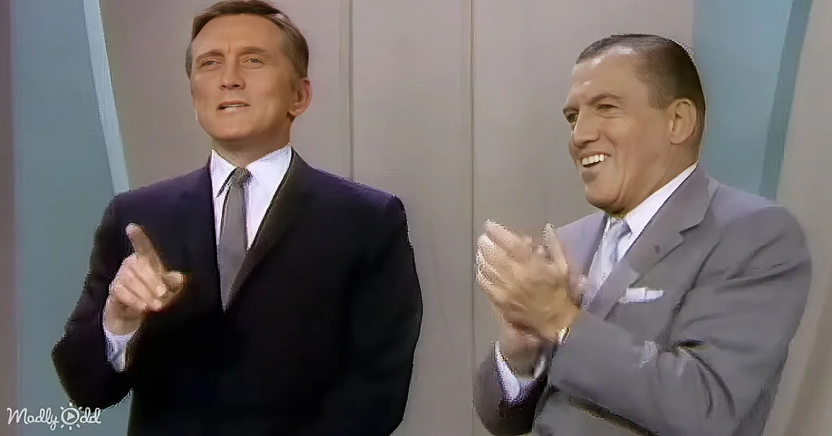 A man in a dark suit points while another in a light suit claps, both smiling.