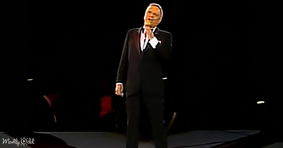 A man in a black suit stands on stage, holding a microphone, with a dark background.