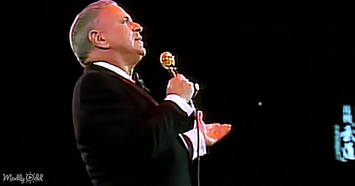 A man in a suit holds a golden award, looking up with a smile, against a dark background.