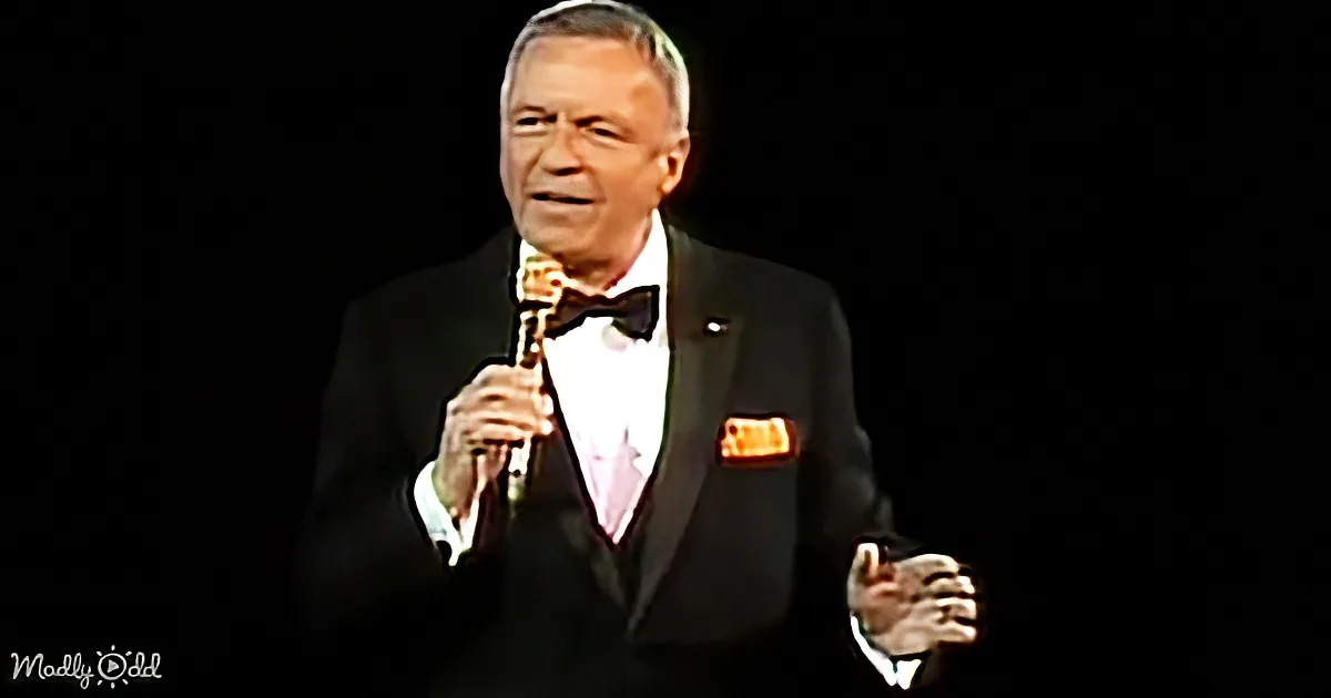 A man in a tuxedo holding a microphone, wearing a bow tie and pocket square, standing against a dark background.