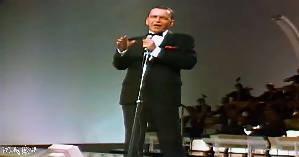 A man in a suit sings into a microphone on stage, with an orchestra in the background.