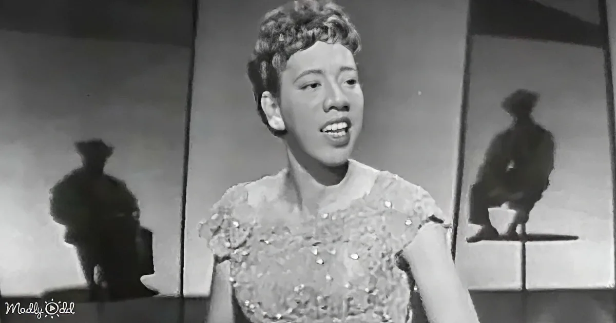 A woman in a beaded dress sings, silhouettes of two figures in the background.