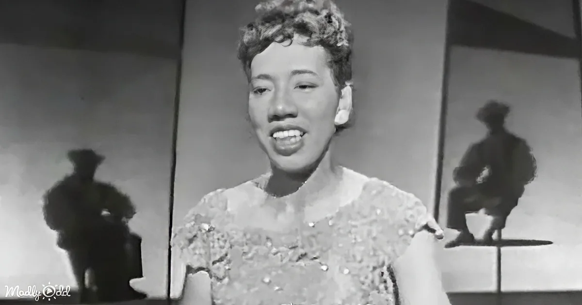 A woman in a patterned dress sings, silhouettes of two figures in the background.