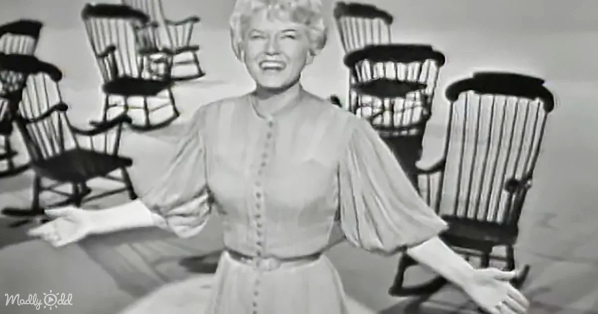 A woman in a vintage dress smiles joyfully with her arms outstretched, surrounded by multiple chairs.