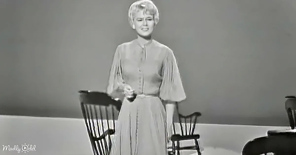 A woman in a vintage dress stands between two chairs, smiling, in a black-and-white setting.