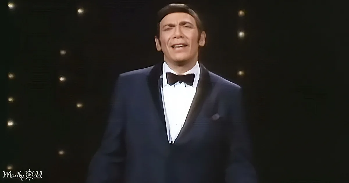 A man in a tuxedo sings on stage with a dark background and small lights.