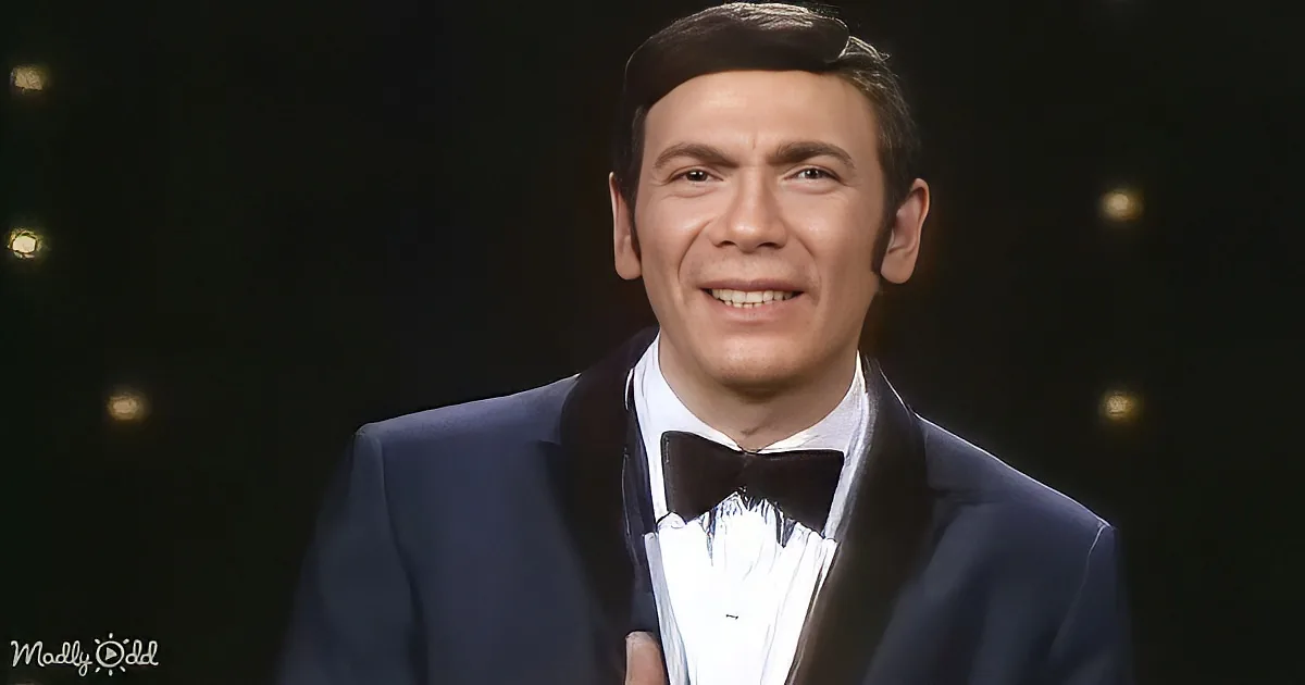 A man in a tuxedo smiles, wearing a bow tie, against a dark background with small lights.