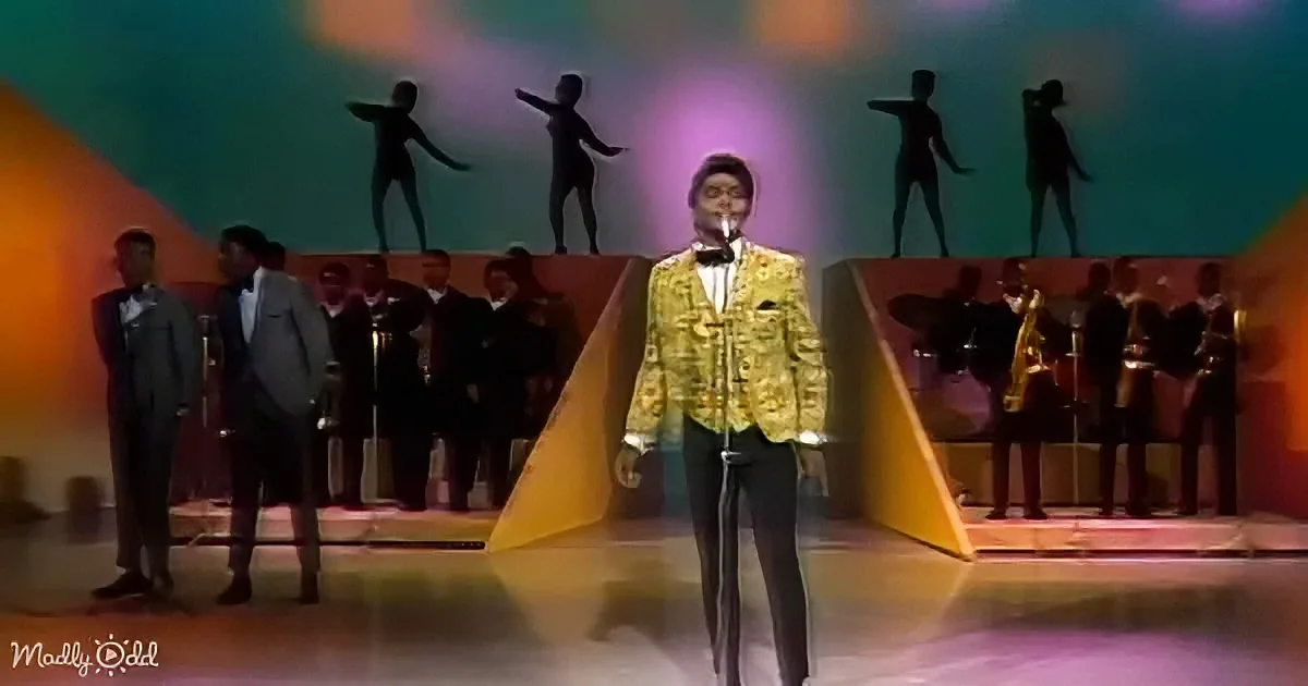 A man in a gold jacket sings on stage with a band and dancers in the background.