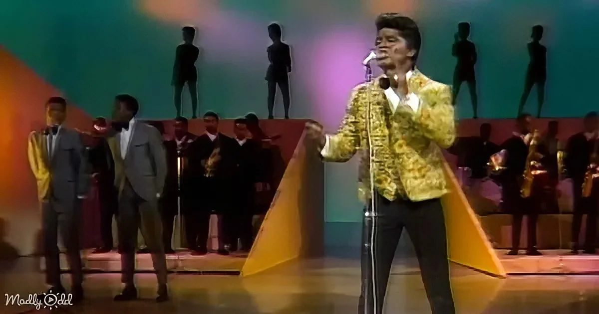 A man in a gold jacket sings on stage with a band and dancers behind him.