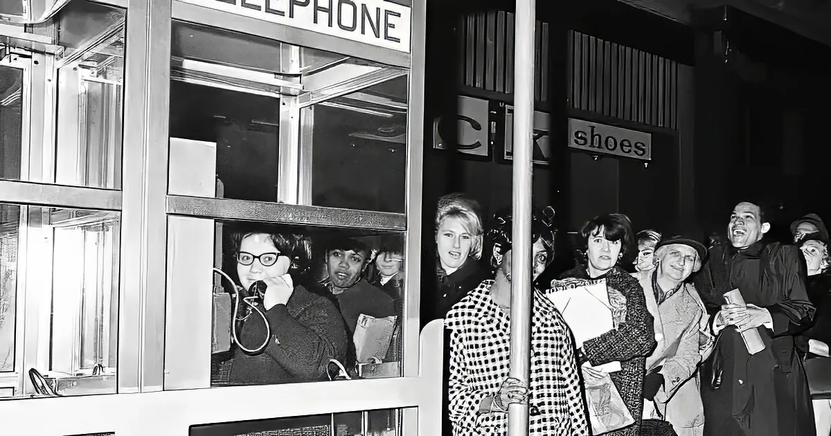 A woman in a phone booth speaks into a microphone as a crowd gathers outside, some holding drinks.