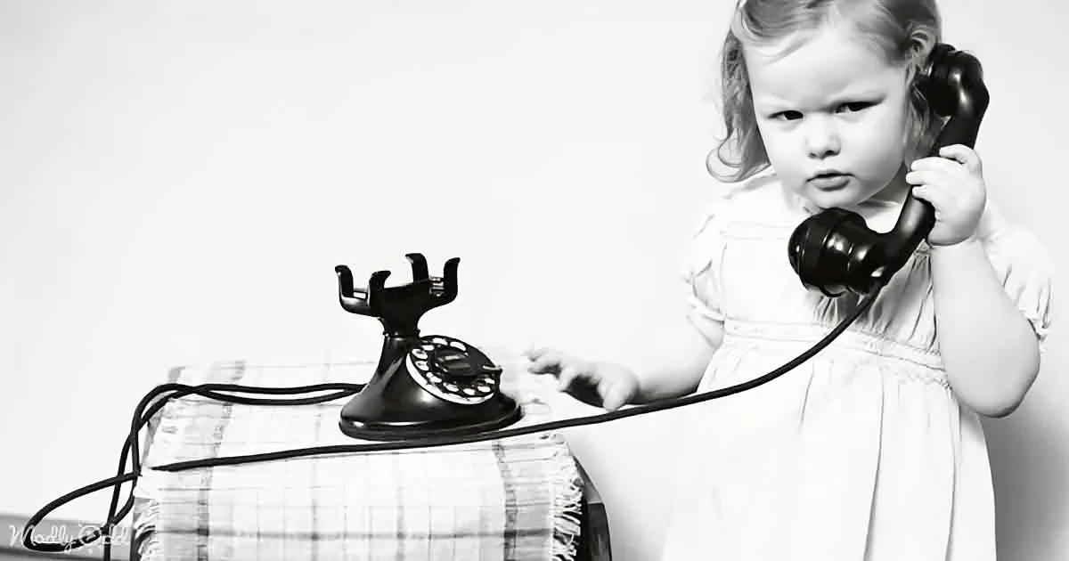 A young child in a white dress holds a vintage telephone receiver, appearing to make a call.