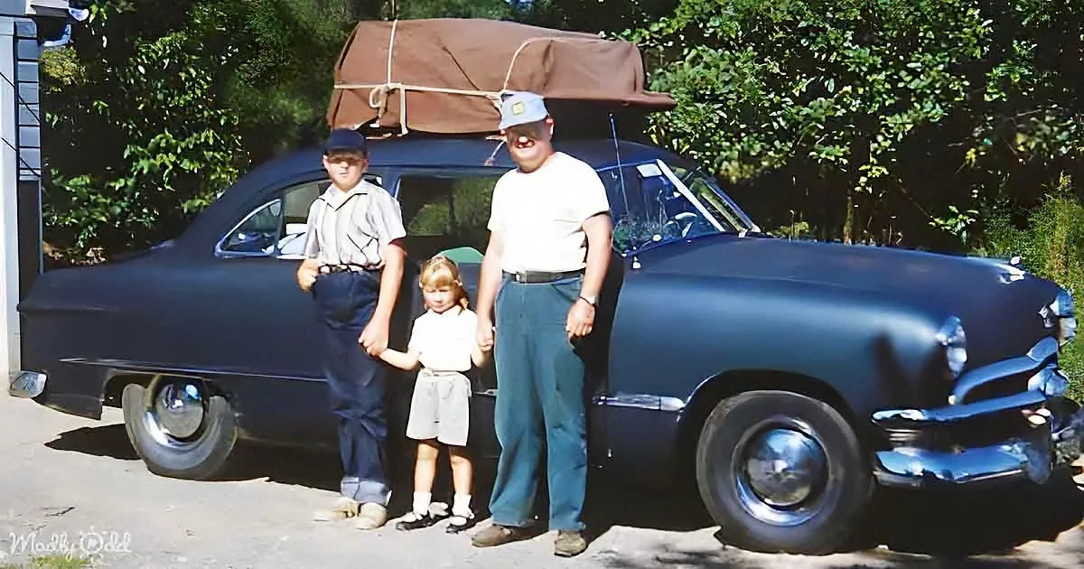 A man, boy, and girl stand in front of a classic car with a brown tarp on the roof.