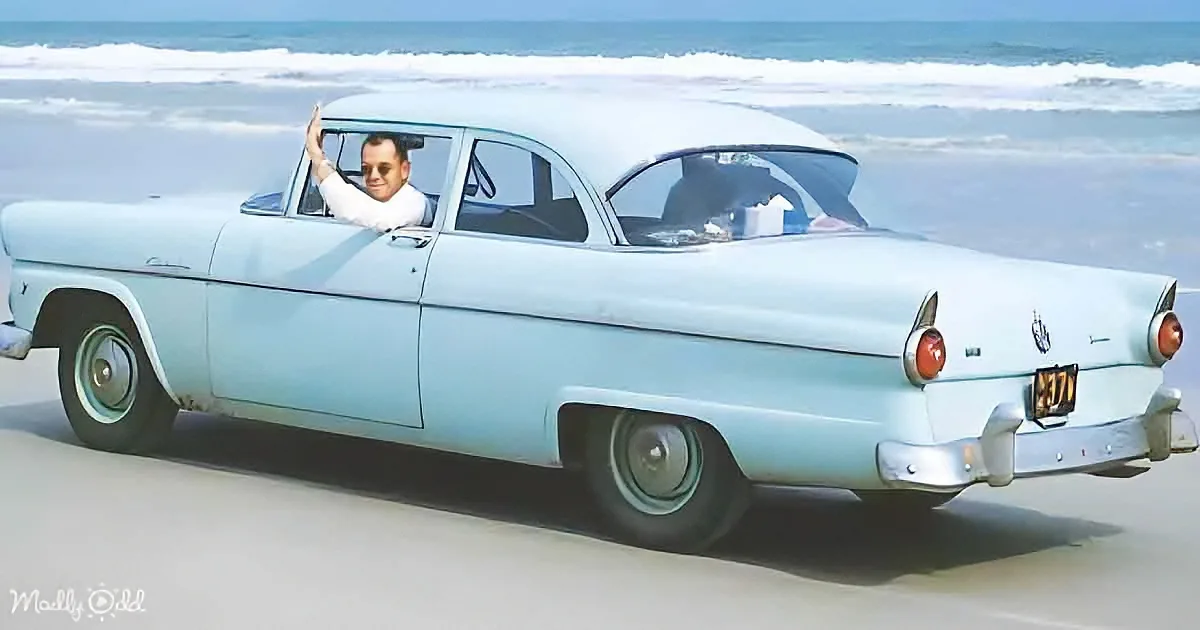 A vintage light blue car drives on a beach, with a person waving from the passenger seat.
