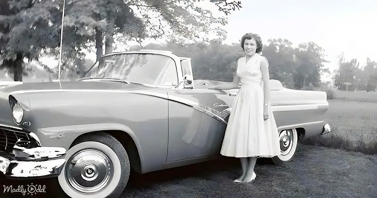 A woman in a white dress stands beside a classic car, smiling, with trees and grass in the background.