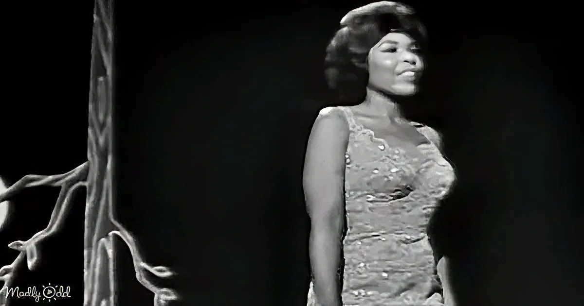 A woman in a lace dress stands on stage, smiling, with a dark background and a decorative column.