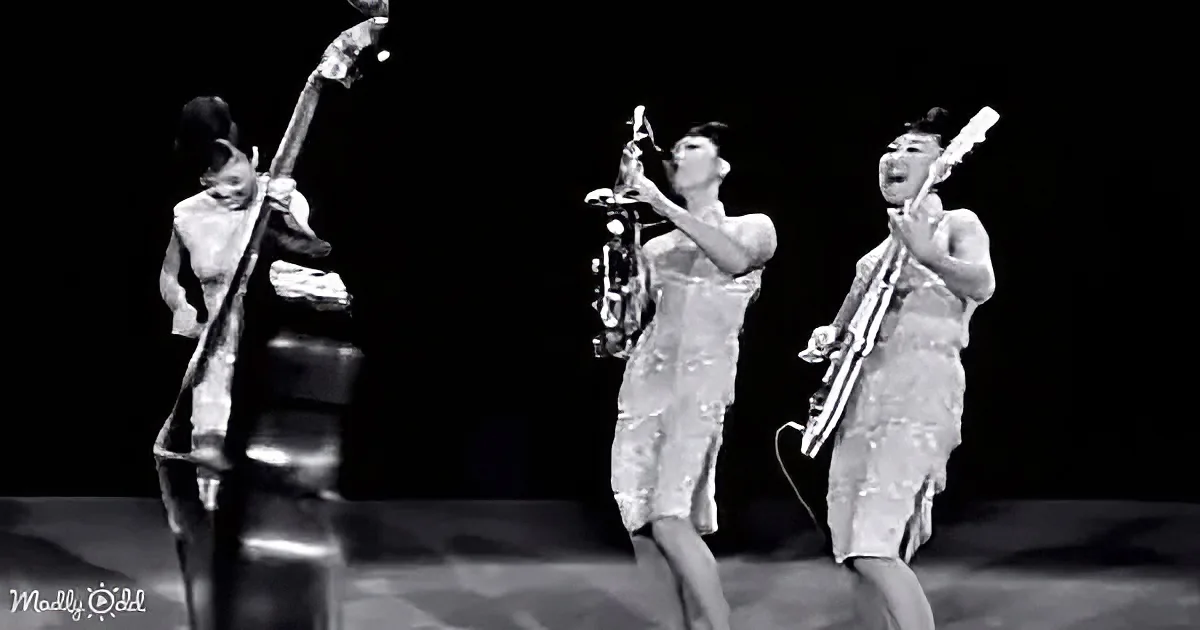 A musician in a silver outfit plays a saxophone on stage, accompanied by others with a double bass and another instrument.