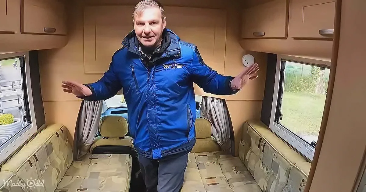 A man in a blue jacket stands inside a camper van with his arms outstretched, showcasing the interior.