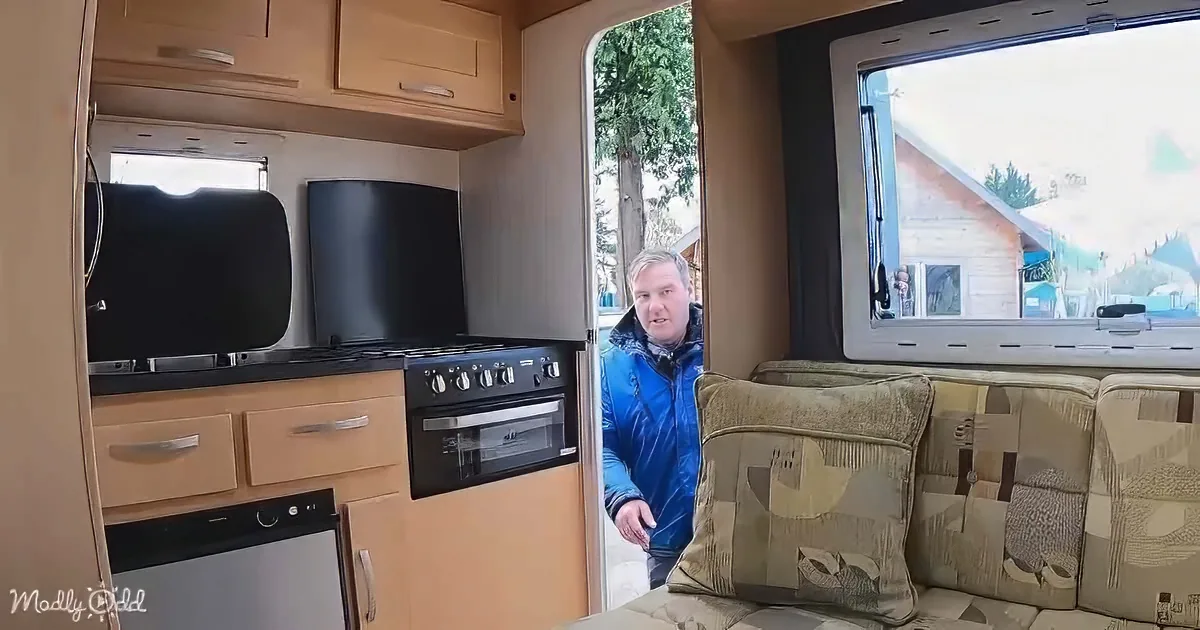 A man in a blue jacket stands in a cozy, compact RV interior with a kitchen and seating area.