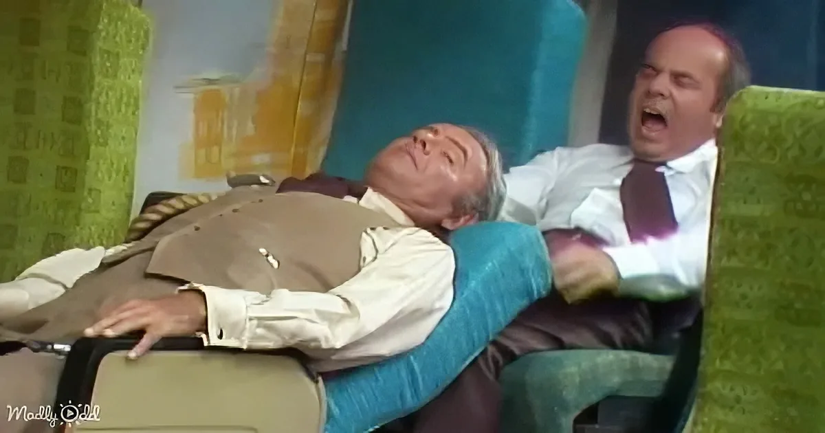 A man in a suit is lying on a chair, appearing to be asleep or unconscious, with another man in a white shirt and tie...