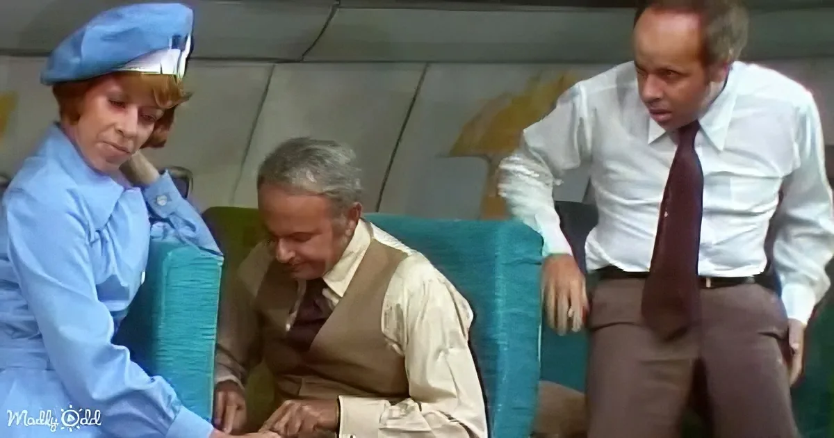 A man in a vest is seated, leaning forward, with a flight attendant and another man observing him.