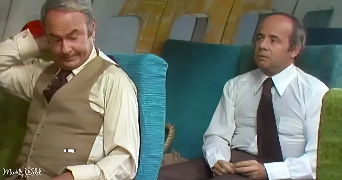 Two men in formal attire sit on a train, one leaning back, the other looking ahead, with teal seats and wooden paneling...