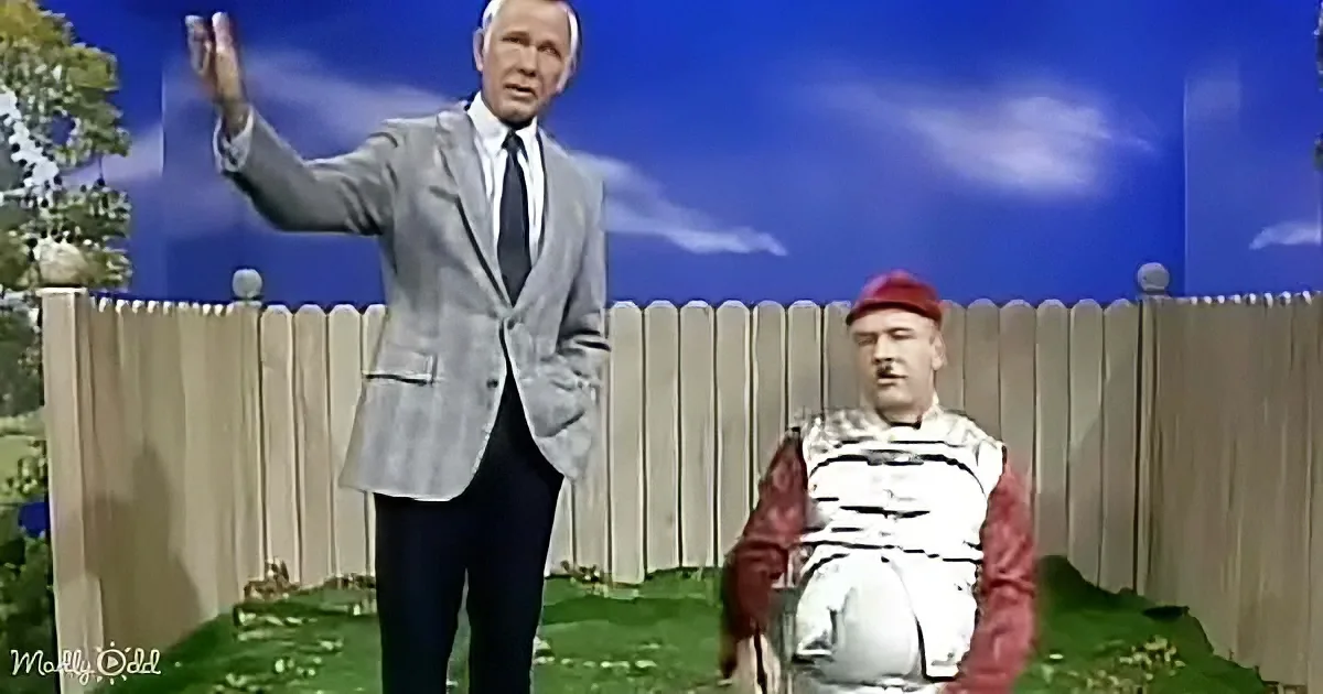 A man in a suit gestures dramatically while another in medieval attire sits on a stool in a grassy yard.
