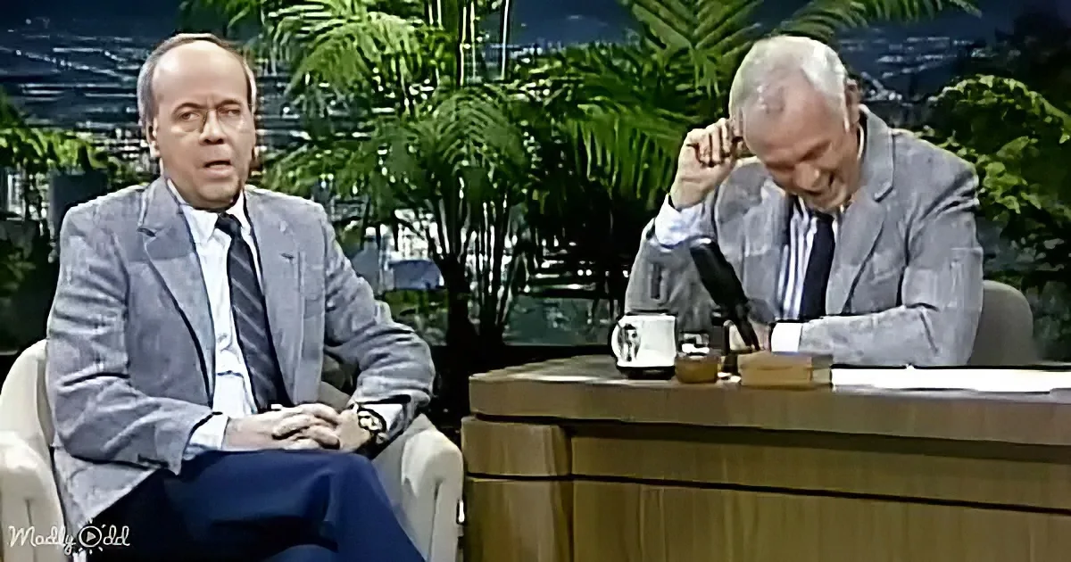 A man in a suit sits in a chair, while another man leans forward at a desk with a microphone, in a studio setting.