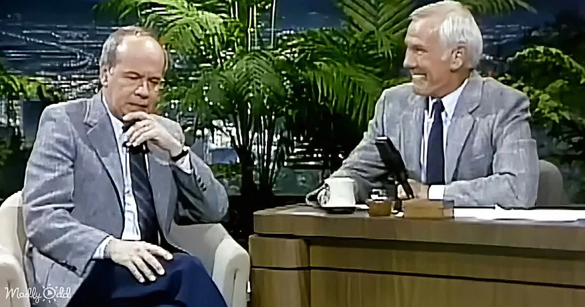 Two men in suits sit on a talk show set, one smiling, the other thoughtful, with tropical plants in the background.